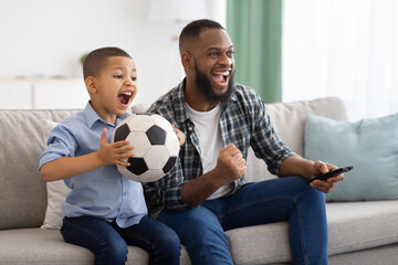 Joyful African Dad And Son Watching Sports On TV Indoors