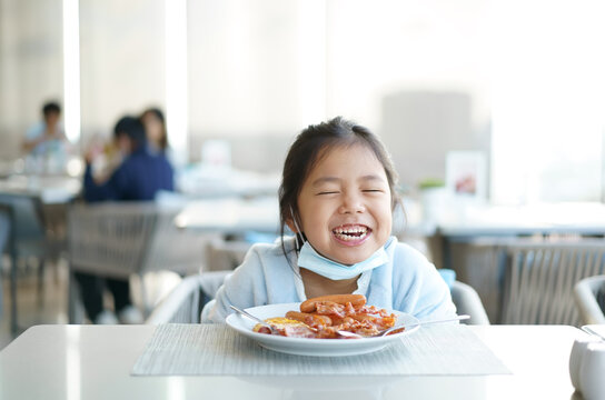 Asian Child Smile Or Kid Girl Wearing Face Mask Under Chin To Enjoy Eating Delicious Food By Sausage Bacon On White Dish With Happy Laugh For Lunch Or Breakfast In Morning At Restaurant Or Food Court