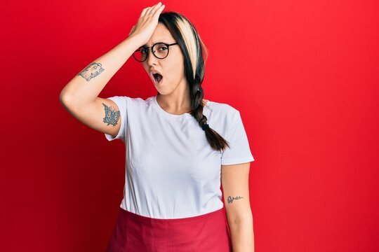 Young hispanic woman wearing professional waitress apron surprised with hand on head for mistake, remember error. forgot, bad memory concept.