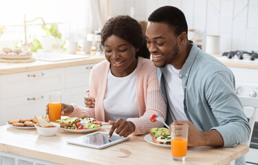 Happy Black Couple Using Digital Tablet While Having Breakfast In Kitchen