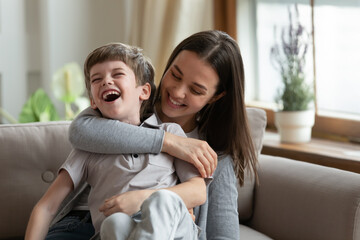 Overjoyed young Caucasian mom and small 7s son have fun play together on weekend in living room. Happy mother feel playful engaged in funny game tickle little boy child. Family entertainment concept. - obrazy, fototapety, plakaty
