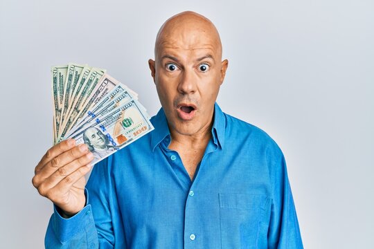 Middle age bald man holding dollars scared and amazed with open mouth for surprise, disbelief face