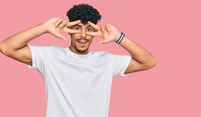 Young arab man wearing casual white t shirt doing peace symbol with fingers over face, smiling cheerful showing victory