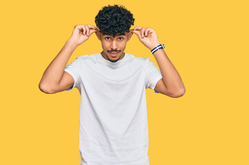 Young arab man wearing casual white t shirt smiling pulling ears with fingers, funny gesture....