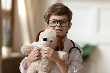 Headshot portrait of little 7s boy child in glasses and medical white uniform hold fluffy toy bear play clinic game. Happy small kid have fun act as future doctor or vet. Healthcare, medicine concept. - obrazy, fototapety, plakaty