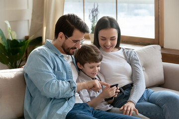 Smiling young Caucasian parents relax on sofa at home with small preschooler son teach using modern cellphone. Happy family with little 6s boy child have fun browsing smartphone. Technology concept.