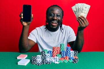 Handsome young black man playing poker holding smartphone and dollars smiling and laughing hard out...