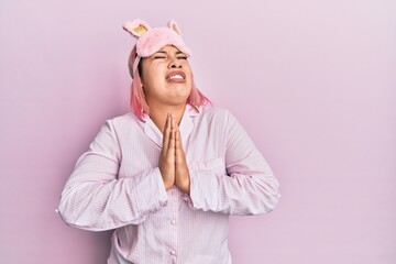 Hispanic woman with pink hair wearing sleep mask and pajama begging and praying with hands together with hope expression on face very emotional and worried. begging.