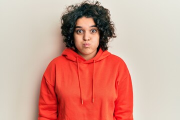 Obraz na płótnie Canvas Young hispanic woman with curly hair wearing casual sweatshirt puffing cheeks with funny face. mouth inflated with air, crazy expression.