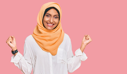 Young brunette woman wearing traditional islamic hijab scarf very happy and excited doing winner gesture with arms raised, smiling and screaming for success. celebration concept.