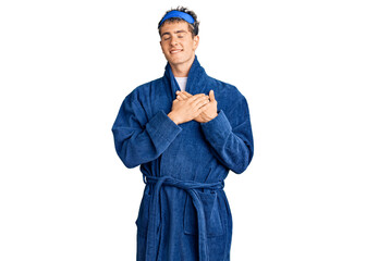 Young handsome man wearing sleep mask and bathrobe smiling with hands on chest with closed eyes and grateful gesture on face. health concept.