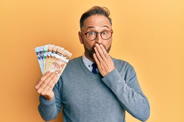 Handsome middle age business man holding canadian dollars covering mouth with hand, shocked and...