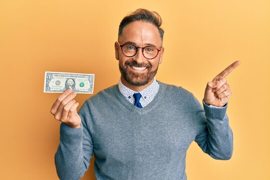 Handsome middle age man holding 1 dollar banknote smiling happy pointing with hand and finger to the side