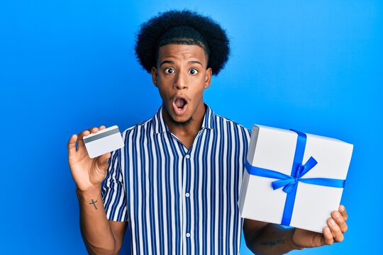 African american man with afro hair holding gift and credit card afraid and shocked with surprise and amazed expression, fear and excited face.