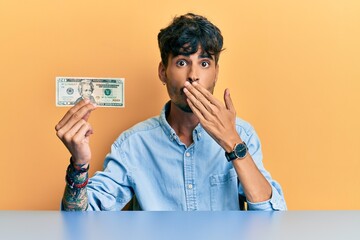 Young hispanic man holding  20 dollar banknote covering mouth with hand, shocked and afraid for mistake. surprised expression