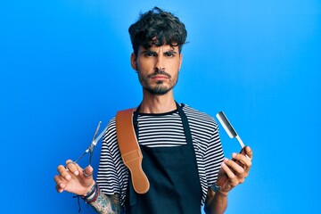 Young hispanic man wearing barber apron holding razor and scissors skeptic and nervous, frowning...