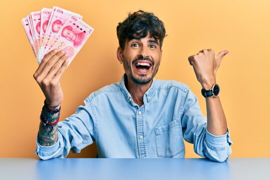 Young hispanic man holding yuan chinese banknotes sitting on the table pointing thumb up to the side smiling happy with open mouth