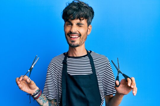 Young hispanic man wearing barber apron holding scissors winking looking at the camera with sexy expression, cheerful and happy face.