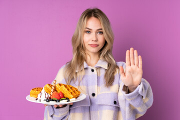 Teenager Russian girl holding waffles isolated on purple background making stop gesture