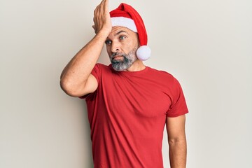 Middle age handsome man wearing christmas hat and summer t-shirt surprised with hand on head for mistake, remember error. forgot, bad memory concept.
