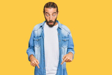 Attractive man with long hair and beard wearing casual denim jacket pointing down with fingers showing advertisement, surprised face and open mouth