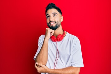 Young man with beard listening to music using headphones with hand on chin thinking about question, pensive expression. smiling and thoughtful face. doubt concept.