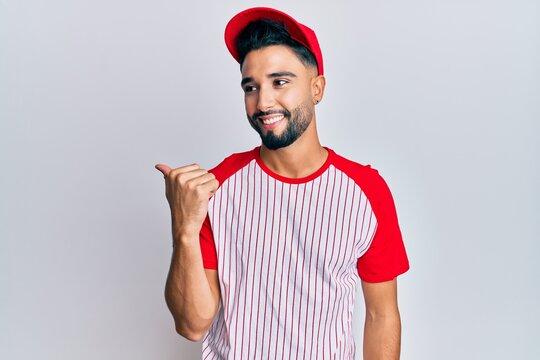 Young man with beard wearing baseball uniform pointing thumb up to the side smiling happy with open mouth