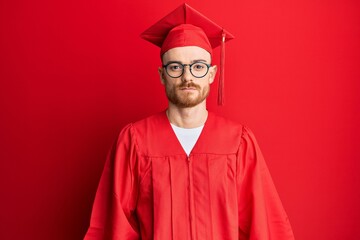 Young redhead man wearing red graduation cap and ceremony robe relaxed with serious expression on face. simple and natural looking at the camera.