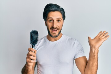 Young hispanic man styling hair using comb celebrating victory with happy smile and winner expression with raised hands