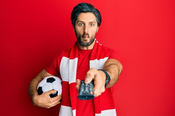 Young hispanic man football hooligan holding ball using tv control making fish face with mouth and squinting eyes, crazy and comical.