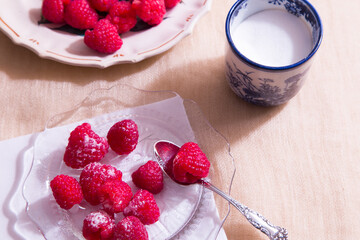 High angle selective focus view of raspberries sprinkled with sugar on clear glass plate, with other fruits on plate in soft focus background