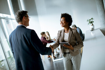 Businesswoman holding digital tablet and looking at handsome colleague while shaking hands in office - Powered by Adobe