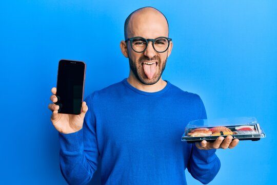 Young hispanic man holding take away food showing smartphone screen sticking tongue out happy with funny expression.