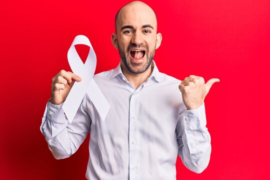 Young handsome bald man holding white cancer ribbon pointing thumb up to the side smiling happy with open mouth