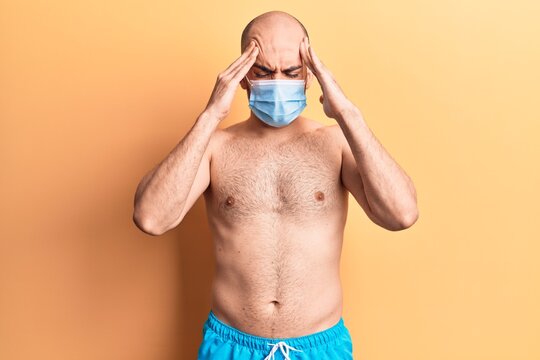 Young handsome bald man shirtless wearing swimwear and medical mask with hand on head, headache because stress. suffering migraine.
