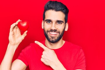 Young handsome man with beard holding red heart smiling happy pointing with hand and finger