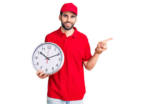 Young handsome man with beard wearing delivery uniform holding big clock smiling happy pointing with hand and finger to the side