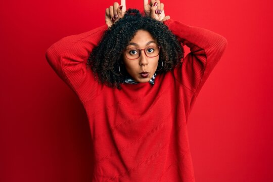 Beautiful african american woman with afro hair wearing sweater and glasses doing funny gesture with finger over head as bull horns