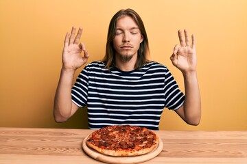 Handsome caucasian man with long hair eating tasty pepperoni pizza relax and smiling with eyes closed doing meditation gesture with fingers. yoga concept.