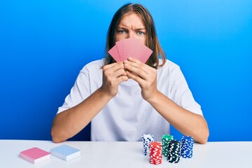 Handsome caucasian man with long hair playing gambling poker covering face with cards skeptic and nervous, frowning upset because of problem. negative person.