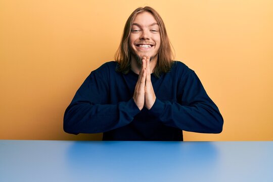Handsome caucasian man with long hair wearing casual clothes sitting on the table praying with hands together asking for forgiveness smiling confident.