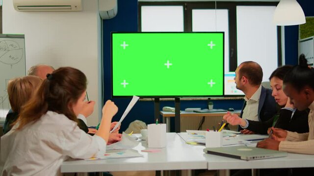 In corporate office meeting room stands green mock-up screen TV or interactive digital whiteboard in horizontal mode. Multiethnical businesspeople working, brainstorming in professional start up