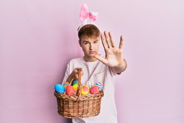 Young caucasian man wearing cute easter bunny ears holding wicker basket with colored eggs with open hand doing stop sign with serious and confident expression, defense gesture