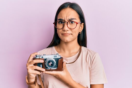 Young asian woman holding vintage camera clueless and confused expression. doubt concept.