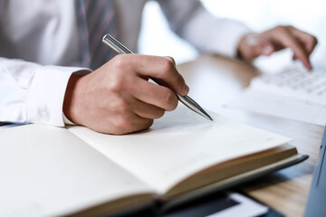 A male employee is taking notes in his notebook, A male employee is using a calculator and taking notes in a notebook.