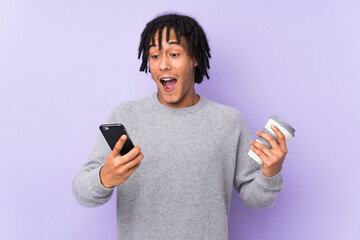 Young african american man isolated on purple background holding coffee to take away and a mobile
