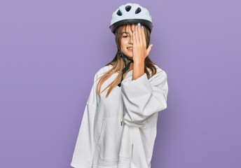 Teenager caucasian girl wearing bike helmet covering one eye with hand, confident smile on face and surprise emotion.