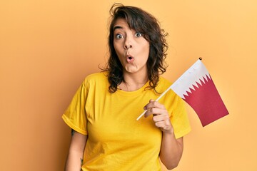 Young hispanic woman holding qatar flag scared and amazed with open mouth for surprise, disbelief face