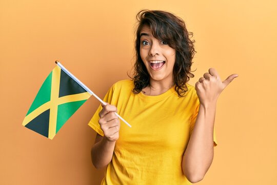 Young hispanic woman holding jamaica flag pointing thumb up to the side smiling happy with open mouth