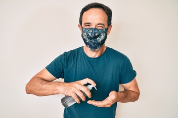 Middle age hispanic man wearing covid-19 protection mask using hand sanitizer gel winking looking...